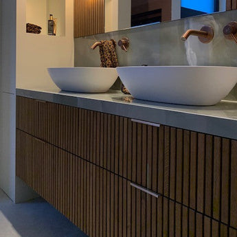 Salle de bains moderne avec meubles de salle de bains en bois à lattes, deux lavabos blancs et robinets cuivrés. Le meuble de salle de bain est équipé de poignées de meuble yessential.fr Enzo Rose Gold, tandis qu'une serviette à imprimé léopard est suspendue au-dessus des lavabos.