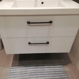 Un meuble sous-vasque de salle de bains moderne Ramiro Black Deluxe de yessential.fr, avec des poignées noires chics, est monté sur un mur carrelé au-dessus d'un tapis à rayures grises et blanches, avec le bord d'un lavabo visible sur le dessus.