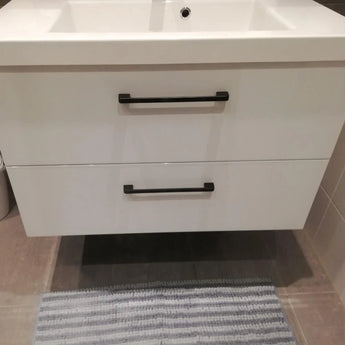 Un meuble sous-vasque de salle de bains moderne Ramiro Black Deluxe de yessential.fr, avec des poignées noires chics, est monté sur un mur carrelé au-dessus d'un tapis à rayures grises et blanches, avec le bord d'un lavabo visible sur le dessus.