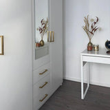 Une chambre moderne et minimaliste avec un mobilier blanc, comprenant l'armoire yessential.fr Vasco Gold avec des poignées dorées et un miroir, ainsi qu'un petit bureau décoré de vases de fleurs séchées et d'un globe sur un sol en bois sombre.