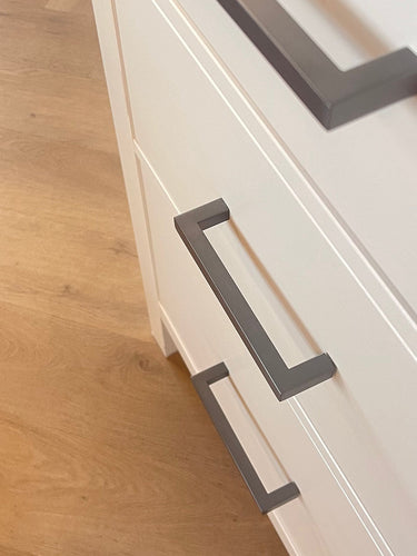 Gros plan d'une armoire blanche avec trois tiroirs, chacun équipé de poignées Vasco Gunmetal de yessential.fr. Placé sur un parquet en bois clair, ce meuble allie style moderne et fonctionnalité pour n'importe quelle pièce.