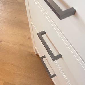 Gros plan d'une armoire blanche avec trois tiroirs, chacun équipé de poignées Vasco Gunmetal de yessential.fr. Placé sur un parquet en bois clair, ce meuble allie style moderne et fonctionnalité pour n'importe quelle pièce.