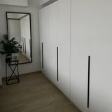 Une chambre moderne avec des armoires yessential.fr Enzo Black avec de longues poignées profilées noires, un grand miroir au sol, une petite table noire avec une plante et des photos encadrées sur un parquet en bois clair.