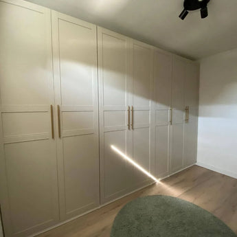 Un ensemble de grandes armoires beiges yessential.fr Remo Gold entourent un mur d'une pièce avec du parquet en bois clair et un tapis circulaire vert clair, tandis que la lumière du soleil projette une diagonale lumineuse sur les portes de l'armoire.