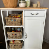 Une armoire de cuisine blanche avec un dessus en bois et une poignée de meuble classique de yessential.fr présente trois paniers en osier contenant des casseroles et de la nourriture. Sur le dessus se trouvent le Daro Ceramic Black, deux petites carafes, un bocal en verre, un bol en bois et une boîte. Un tiroir et un placard sont visibles.