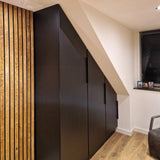 Le meuble Enzo Black de yessential.fr, avec poignées d'angle noires, s'intègre parfaitement sous un plafond mansardé dans une pièce moderne avec des boiseries, du parquet clair et une petite fenêtre avec stores.