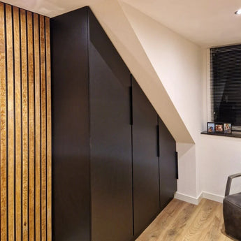 Le meuble Enzo Black de yessential.fr, avec poignées d'angle noires, s'intègre parfaitement sous un plafond mansardé dans une pièce moderne avec des boiseries, du parquet clair et une petite fenêtre avec stores.