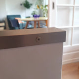 Gros plan d'une vis Enzo en acier inoxydable de yessential.fr fixée sur une surface blanche, avec un arrière-plan flou montrant une table, des chaises et des plantes d'intérieur dans une pièce lumineuse.