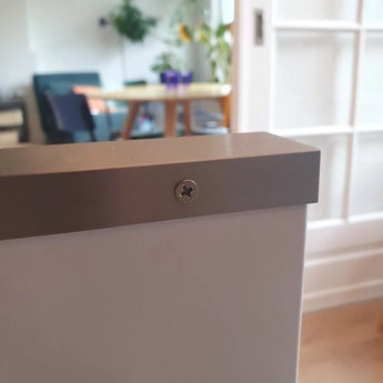 Gros plan d'une vis Enzo en acier inoxydable de yessential.fr fixée sur une surface blanche, avec un arrière-plan flou montrant une table, des chaises et des plantes d'intérieur dans une pièce lumineuse.