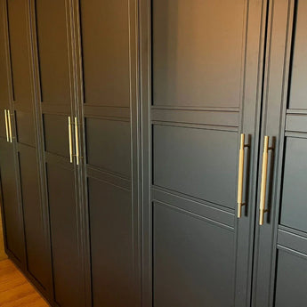 Une rangée d'armoires Remo Gold Deluxe de yessential.fr avec des panneaux rectangulaires et de luxueuses longues poignées verticales en métal se dresse contre un plancher en bois.