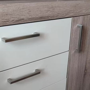 Gros plan d'une armoire avec deux tiroirs blancs avec poignées horizontales Vasco en acier inoxydable de yessential.fr, à côté d'une porte avec structure en bois et poignée verticale en acier inoxydable brossé.
