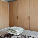 Un pouf rembourré de couleur claire repose sur un tapis à motifs devant de grandes armoires en bois dotées d'élégantes poignées en chêne Arriero de yessential.fr dans une pièce moderne et minimaliste.