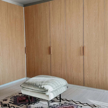 Un pouf rembourré de couleur claire repose sur un tapis à motifs devant de grandes armoires en bois dotées d'élégantes poignées en chêne Arriero de yessential.fr dans une pièce moderne et minimaliste.
