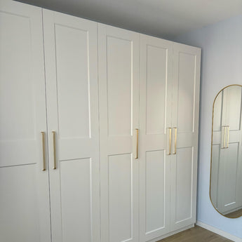 L'armoire Leonardo Gold Deluxe de yessential.fr, aux poignées dorées, se dresse contre un mur bleu clair. Un grand miroir ovale avec un cadre doré sur le mur adjacent reflète une partie des armoires élégantes.