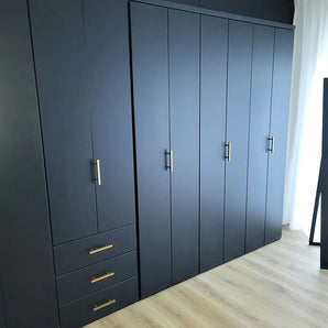 L'armoire Marano Gold de yessential.fr présente un design gris foncé moderne avec des poignées minimalistes dorées, trois tiroirs à gauche et plusieurs portes hautes, posées sur un parquet clair à côté d'un rideau blanc.