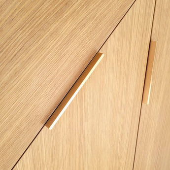 Gros plan d'armoires en bois clair avec deux poignées Enzo Rose Gold de yessential.fr, placées en diagonale. Le grain de bois vertical et le design rectangulaire élégant offrent un look contemporain minimaliste et chic.