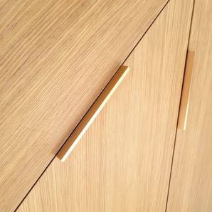 Gros plan d'armoires en bois clair avec deux poignées Enzo Rose Gold de yessential.fr, placées en diagonale. Le grain de bois vertical et le design rectangulaire élégant offrent un look contemporain minimaliste et chic.
