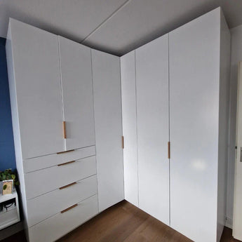 L'armoire d'angle Enzo Rose Gold de yessential.fr possède des portes blanc brillant, des poignées modernes, quatre tiroirs à gauche, des armoires hautes des deux côtés et se dresse élégamment sur un plancher en bois dans une pièce moderne.