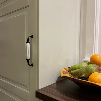 Dans un coin cuisine se trouve un placard de couleur crème avec la poignée Daro Ceramic Black yessential.fr et un bol de poires, une pomme et des oranges sur un plan de travail en bois foncé.
