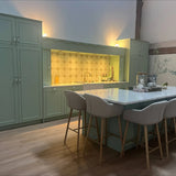 Une cuisine moderne avec des armoires vert sauge avec des boutons d'armoire Daro Ceramic Antique Gold de yessential.fr, un îlot en marbre avec quatre tabourets de bar rembourrés beiges, du parquet et un dosseret de carreaux hexagonaux sous un éclairage chaleureux.