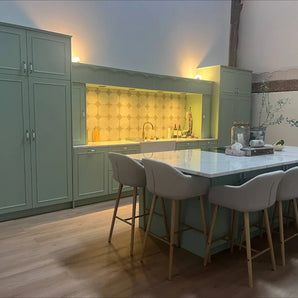 Une cuisine moderne avec des armoires vert sauge avec des boutons d'armoire Daro Ceramic Antique Gold de yessential.fr, un îlot en marbre avec quatre tabourets de bar rembourrés beiges, du parquet et un dosseret de carreaux hexagonaux sous un éclairage chaleureux.