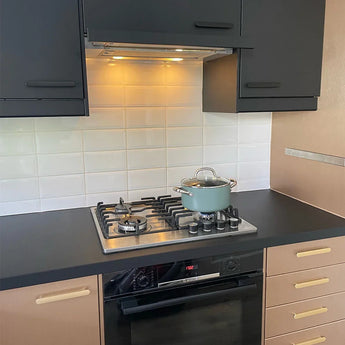Une cuisine moderne comprend des armoires Arriero Black Deluxe noires et beiges de yessential.fr, des poignées de luxe, une cuisinière à gaz avec une poêle bleue et un couvercle en verre, un dosseret en carrelage blanc et un four sous la cuisinière.