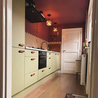 Une cuisine américaine moderne avec des armoires vert clair, des poignées yessential.fr Arriero Walnut, du parquet, une crédence beige festonnée, des murs et un plafond rouge foncé, des lampes suspendues et une porte blanche au bout. La lumière naturelle entre par une fenêtre sur la droite.
