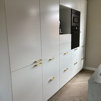 Cuisine moderne avec armoires blanches, poignées Vasco Gold de yessential.fr, four encastré noir et plancher à chevrons en bois clair. Les murs neutres et la décoration minimaliste créent un look épuré et élégant.