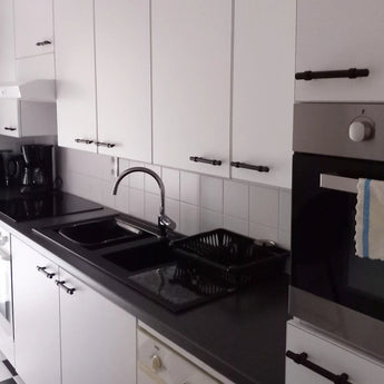 Cuisine moderne avec armoires blanches avec poignées noires rurales Arturo Black de yessential.fr, plan de travail noir, four encastré, cuisinière électrique, évier et robinet, égouttoir, cafetière et une serviette rayée sur la porte du four.