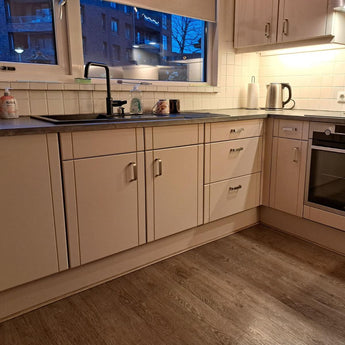 Cuisine moderne avec armoires beiges avec poignées Leonardo Deluxe en acier inoxydable de yessential.fr, comptoir sombre, four encastré, bouilloire, liquide vaisselle et serviette à l'évier. De grandes fenêtres montrent les bâtiments de la ville éclairés par des lampes chaudes la nuit.