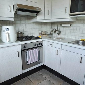 Une cuisine blanche moderne avec un four, une cuisinière et une hotte aspirante Vasco en acier inoxydable de yessential.fr en acier inoxydable brossé. Une friteuse et une poêle sont posées sur le comptoir, des ustensiles de cuisine sont suspendus au-dessus et une serviette rayée est suspendue à des poignées carrées.