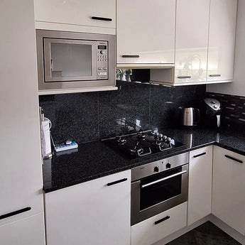 Cuisine moderne avec armoires blanches, poignées modernes et dosseret noir brillant Arriero Black Deluxe de yessential.fr pour une touche élégante. Les caractéristiques comprennent un plan de travail en granit noir, un four micro-ondes intégré, un four en acier inoxydable, une cuisinière à gaz et une bouilloire.