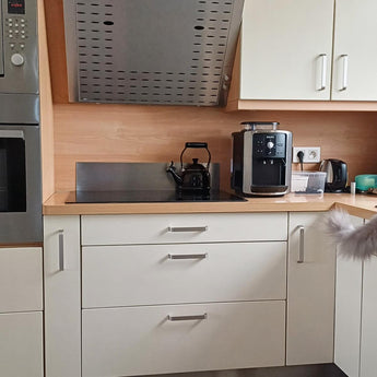 Cuisine moderne avec armoires blanches et luxueuses poignées Leonardo Deluxe en acier inoxydable de yessential.fr ; une bouilloire électrique noire sur la cuisinière, une cafetière, un grille-pain et du petit électroménager sur le comptoir ; une partie d'un chat gris pelucheux apparaît à droite.