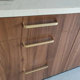 Un gros plan de trois tiroirs en bois avec la poignée de cuisine de luxe Leonardo Gold Deluxe de yessential.fr en or, finis avec un plan de travail blanc. Le bois présente un motif de grain naturel.