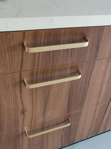 Un gros plan de trois tiroirs en bois avec la poignée de cuisine de luxe Leonardo Gold Deluxe de yessential.fr en or, finis avec un plan de travail blanc. Le bois présente un motif de grain naturel.