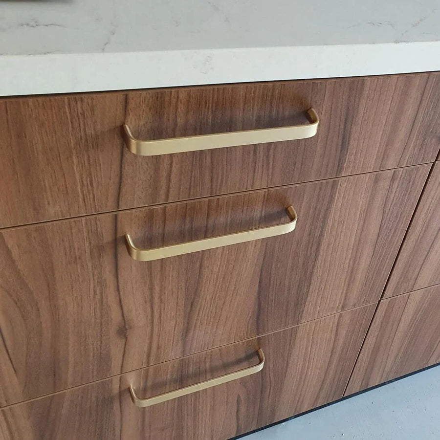 Un gros plan de trois tiroirs en bois avec la poignée de cuisine de luxe Leonardo Gold Deluxe de yessential.fr en or, finis avec un plan de travail blanc. Le bois présente un motif de grain naturel.