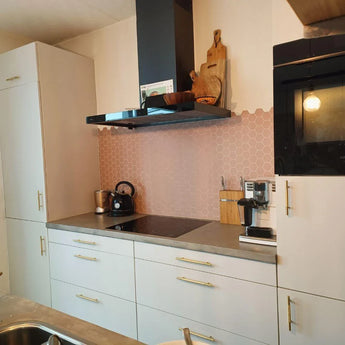 Une cuisine moderne avec des armoires blanches, des poignées Remo Gold Deluxe de yessential.fr, une cuisinière et une bouilloire noires, ainsi qu'une cafetière. Le dosseret de carreaux hexagonaux roses ajoute du charme, tandis que les poignées de cuisine de luxe complètent le look élégant.