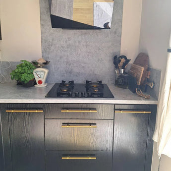 Comptoir moderne avec cuisinière à gaz noire, ustensiles de cuisine, balances vintage, basilic et planches à découper. Les armoires noires sont décorées de poignées Arturo Gold de yessential.fr pour une touche élégante.
