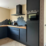 Cuisine moderne avec armoires yessential.fr Enzo Noir mat et poignées d'angle noires, plan de travail en bois, mur arrière en carrelage noir hexagonal, four encastré, cuisinière à gaz, hotte aspirante, plus une petite plante verte sur le plan de travail.