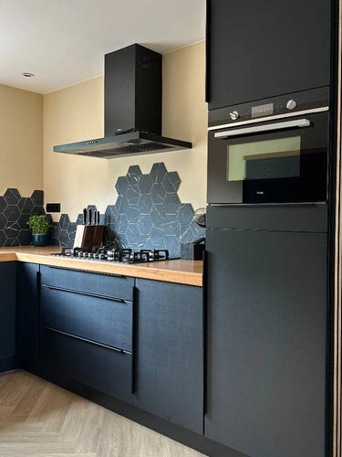 Cuisine moderne avec armoires yessential.fr Enzo Noir mat et poignées d'angle noires, plan de travail en bois, mur arrière en carrelage noir hexagonal, four encastré, cuisinière à gaz, hotte aspirante, plus une petite plante verte sur le plan de travail.