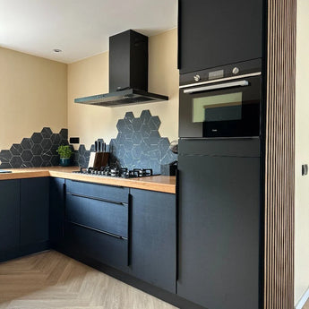 Cuisine moderne avec armoires yessential.fr Enzo Noir mat et poignées d'angle noires, plan de travail en bois, mur arrière en carrelage noir hexagonal, four encastré, cuisinière à gaz, hotte aspirante, plus une petite plante verte sur le plan de travail.