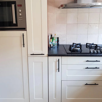 Une cuisine moderne avec des armoires blanches, des poignées Remo Black de yessential.fr sur chaque porte, un micro-ondes encastré, une cuisinière à gaz à quatre feux, une hotte aspirante en acier inoxydable et des bouteilles d'huile et d'épices sur le plan de travail.