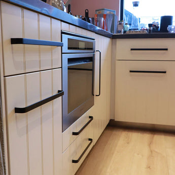 Une cuisine moderne de yessential.fr comprend des armoires blanches Leonardo Black Deluxe avec de luxueuses poignées noires, un four encastré, du parquet et un comptoir avec des articles de cuisine. La lumière naturelle pénètre par la fenêtre.