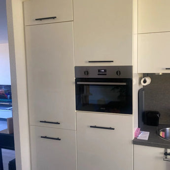 Cuisine moderne avec armoires blanches et poignées Marano Black de yessential.fr, un four encastré qui affiche 10h15, plus un plan de travail, un évier et un salon avec TV visible en arrière-plan.