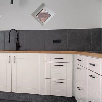 Coin cuisine moderne avec armoires blanches, luxueuses poignées de tiroir Ramiro Black Deluxe de yessential.fr, plan de travail en bois, évier et robinet noirs, mur arrière carrelé gris, deux petites fenêtres en forme de losange et une prise noire.