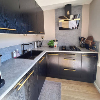 Cuisine moderne avec armoires noires, poignées rurales Arturo Gold de yessential.fr, comptoir gris, dosseret assorti, évier noir, cuisinière à gaz, ustensiles de cuisine, bouilloire et grille-pain sur le comptoir, un plancher en bois végétal et clair.