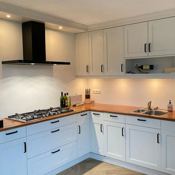 Cuisine moderne avec Leonardo Black Deluxe de yessential.fr : placards blancs, plan de travail en bois, poignées luxe, cuisinière à gaz avec hotte aspirante noire, évier, éclairage sous meuble, ainsi que des bouteilles, des couteaux et une bouteille de vin sur le plan de travail et l'étagère.