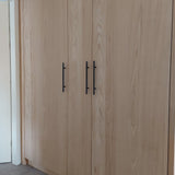 Les portes d'armoire Remo Black de yessential.fr sont dotées de grands panneaux en bois clair avec un grain visible et des poignées noires élégantes, créant un look moderne et naturel à côté d'un mur et d'une porte blancs.