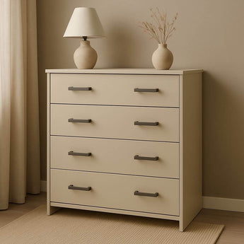 La commode Vasco Gunmetal de yessential.fr dispose de quatre tiroirs avec poignées en bronze et se situe dans une pièce neutre. Au sommet se trouvent une lampe de couleur crème et un vase avec des branches séchées, placés près d'une fenêtre avec des rideaux lumineux.