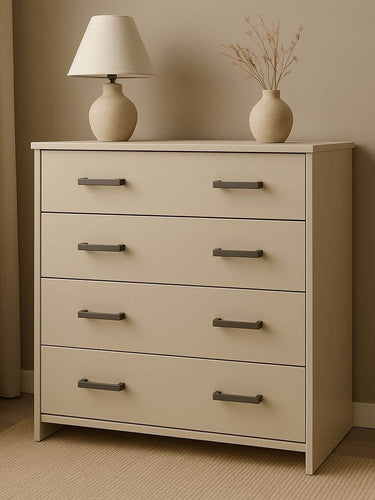 La commode Vasco Gunmetal de yessential.fr dispose de quatre tiroirs avec poignées en bronze et se situe dans une pièce neutre. Au sommet se trouvent une lampe de couleur crème et un vase avec des branches séchées, placés près d'une fenêtre avec des rideaux lumineux.